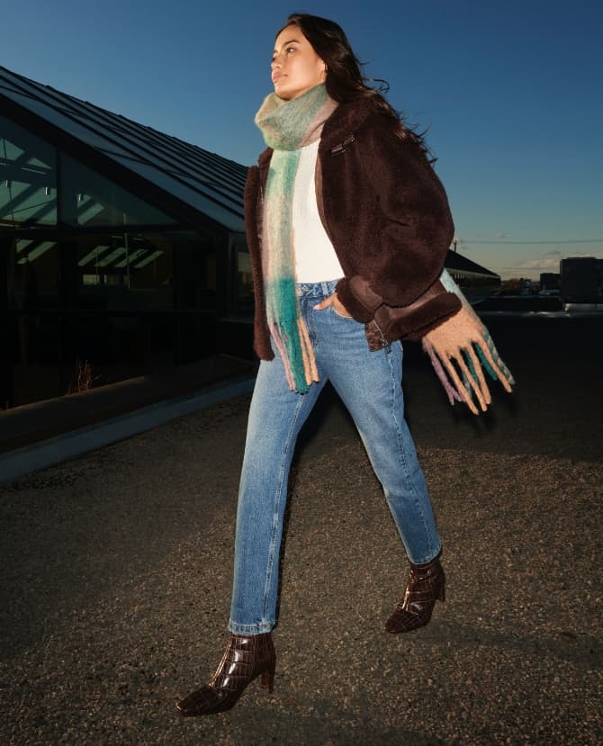 A model wears blue jeans with a white top, sherpa aviator jacket and a madras plaid scarf.