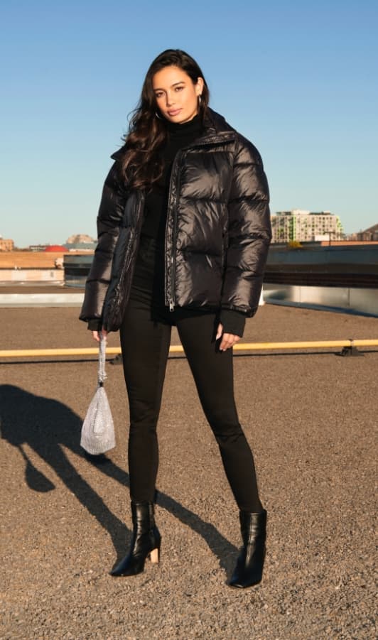 A model wears a black puffer coat with black jeans, and holds a gemmed knotted handbag.